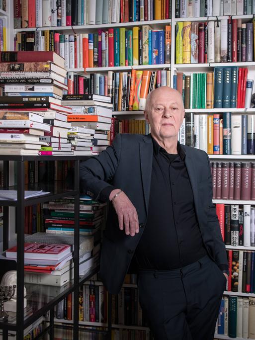 Schriftsteller und Verleger Klaus Bittermann in den Räumen seines Verlags Edition Tiamat in Berlin Kreuzberg. Er steht in einem grauen Anzug vor einem vollen Bücherregal, Buchstapel liegen um ihn herum.