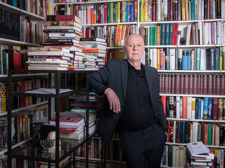 Schriftsteller und Verleger Klaus Bittermann in den Räumen seines Verlags Edition Tiamat in Berlin Kreuzberg. Er steht in einem grauen Anzug vor einem vollen Bücherregal, Buchstapel liegen um ihn herum.