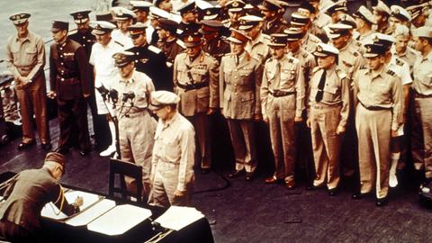 General Umezu Yoshijiro unterzeichnet Dokumente auf einem Schreibtisch zur Kapitulation Japans. Dahinter stehen hochrangige Militärvertreter.