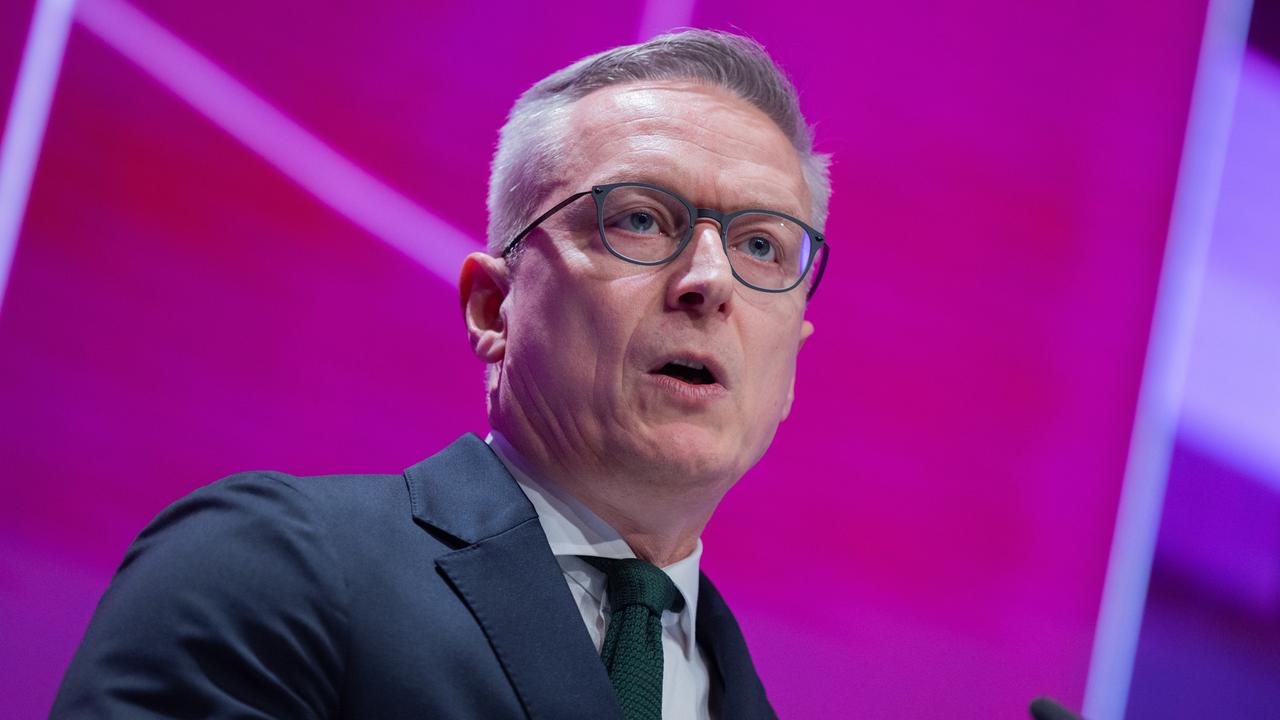 Wildberger steht vor einem violetten Hintergrund und spricht. Er trägt einen dunklen Anzug und Krawatte.