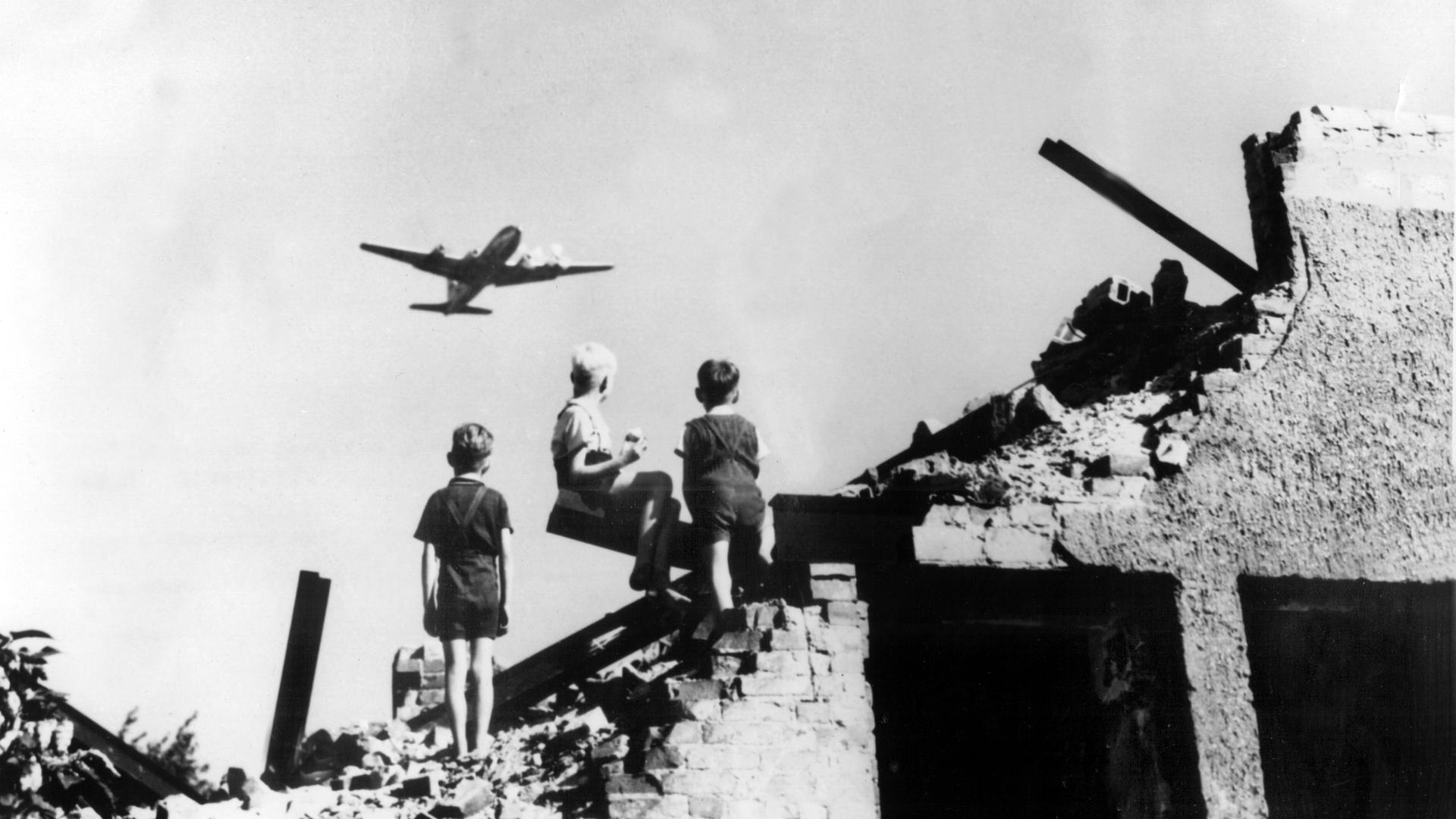 Drei West-Berliner Jungen, die auf einem Trümmerberg spielen, schauen zu einem US-amerikanisches Transportflugzeug empor, das Versorgungsgüter nach West-Berlin bringt. 