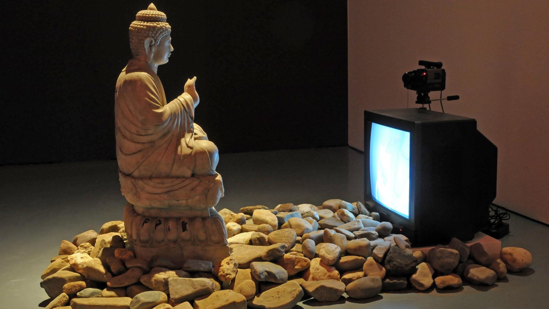 Eine große Buddha-Statue steht in einer Ausstellungshalle. Ihr gegenüber steht ein Röhrenfernseher, davor liegen Steine auf dem Boden. Es handelt sich um die Videoinstallation "Buddha Duchamp Beuys 1989" des koreanischen Medienkünstlers "Nam June Paik" Eine große Buddha-Statue steht in einer Ausstellungshalle. Ihr gegenüber steht ein Röhrenfernseher, davor liegen Steine auf dem Boden. Es handelt sich um die Videoinstallation "Buddha Duchamp Beuys 1989" des koreanischen Medienkünstlers "Nam June Paik"