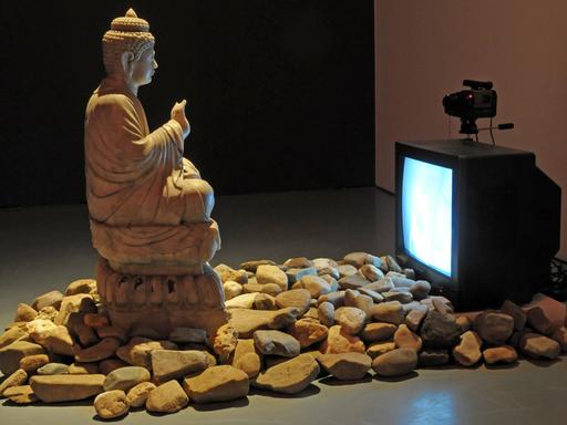 Eine große Buddha-Statue steht in einer Ausstellungshalle. Ihr gegenüber steht ein Röhrenfernseher, davor liegen Steine auf dem Boden. Es handelt sich um die Videoinstallation "Buddha Duchamp Beuys 1989" des koreanischen Medienkünstlers "Nam June Paik"
