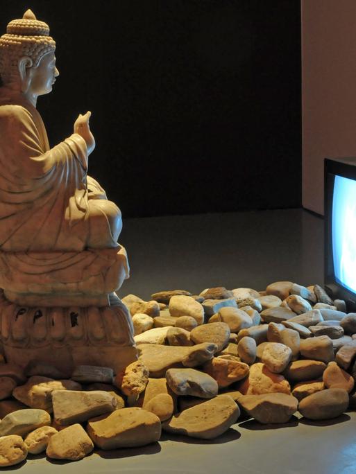 Eine große Buddha-Statue steht in einer Ausstellungshalle. Ihr gegenüber steht ein Röhrenfernseher, davor liegen Steine auf dem Boden. Es handelt sich um die Videoinstallation "Buddha Duchamp Beuys 1989" des koreanischen Medienkünstlers "Nam June Paik"