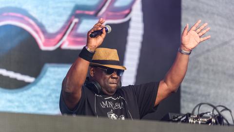 Ein kräftiger Mann steht bei einem Open Air Festival hinter einem Mischpult. Im Hintergrund ist ein buntes Graffiti zu sehen. Er trägt Sonnenbrille und einen Strohhut.