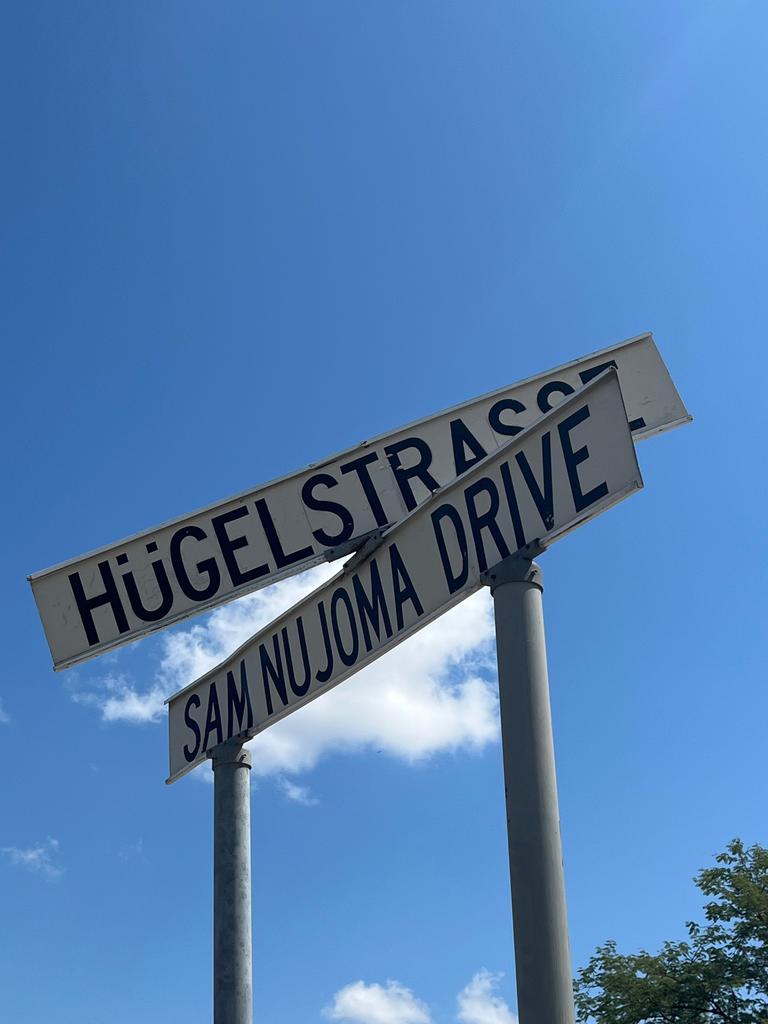 Zwei Schilder markieren eine Kreuzung in Windhoek. Eine Straße ist nach Sam Nujoma benannt, dem Staatsgründer von Namibia, auf dem anderen Schild steht auf Deutsch Hügelstraße