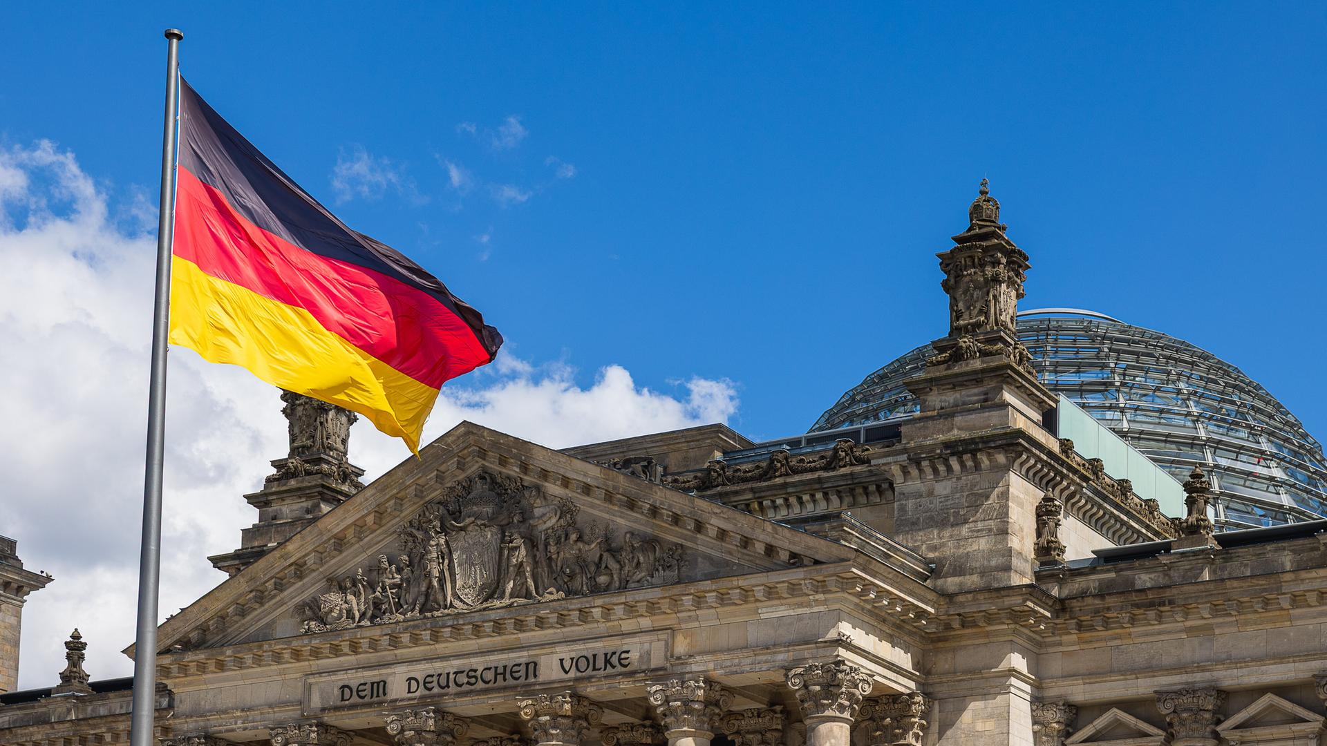 Die deutsche Nationalflagge weht vor dem Reichstagsgebäude in Berlin, Deutschland. Im Hintergrund ist die markante Glaskuppel des Parlamentsgebäudes zu sehen.