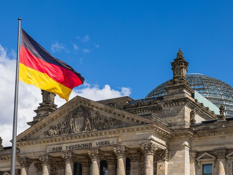 Die deutsche Nationalflagge weht vor dem Reichstagsgebäude in Berlin, Deutschland. Im Hintergrund ist die markante Glaskuppel des Parlamentsgebäudes zu sehen.