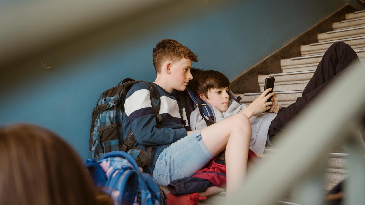 Jungen schauen zusammen auf ein Smartphone während sie auf den Treppen eines Schulgebäudes sitzen.