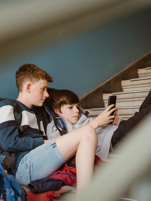 Jungen schauen zusammen auf ein Smartphone während sie auf den Treppen eines Schulgebäudes sitzen. Jungen schauen zusammen auf ein Smartphone während sie auf den Treppen eines Schulgebäudes sitzen.