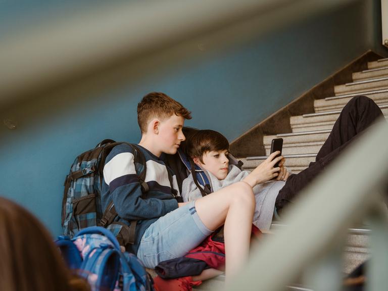 Jungen schauen zusammen auf ein Smartphone während sie auf den Treppen eines Schulgebäudes sitzen. Jungen schauen zusammen auf ein Smartphone während sie auf den Treppen eines Schulgebäudes sitzen.