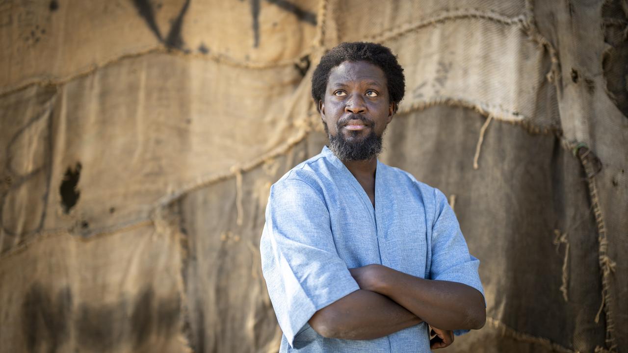 Mahama steht vor einem Kunstwerk aus Jutesäcken. Er trägt ein hellblaues Hemd und verschränkt die Arme.