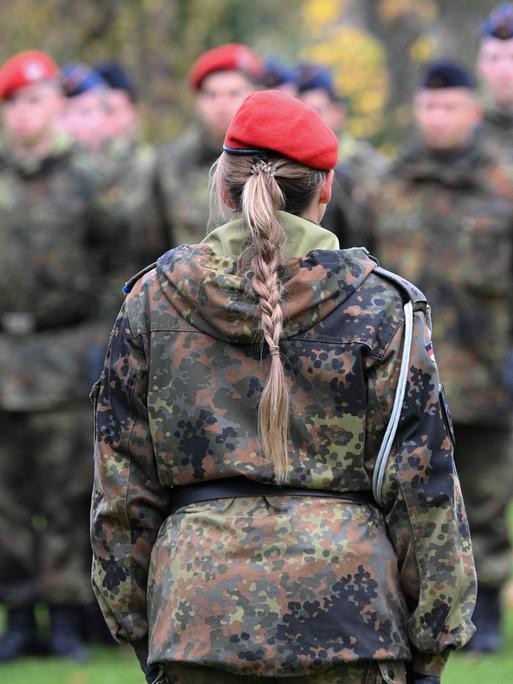 Eine Frau mit blondem Zopf in Bundeswehrkleidung ist von hinten zu sehen, im Hintergrund sind verschwommen männliche Soldaten zu erkennen