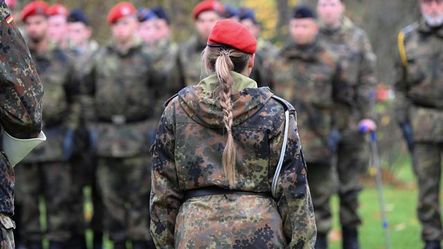 Eine Frau mit blondem Zopf in Bundeswehrkleidung ist von hinten zu sehen, im Hintergrund sind verschwommen männliche Soldaten zu erkennen