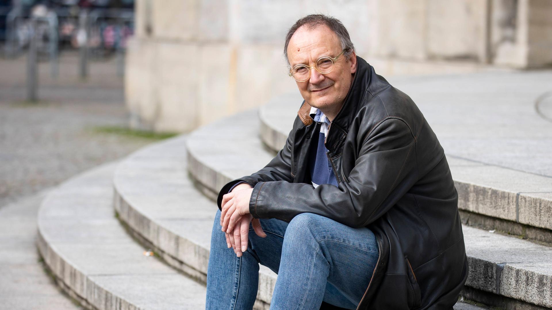 Thomas Brussig sitzt in Lederjacke und Jeans lässig auf den Stufen vor der Berliner Volksbühne und blickt in die Kamera.