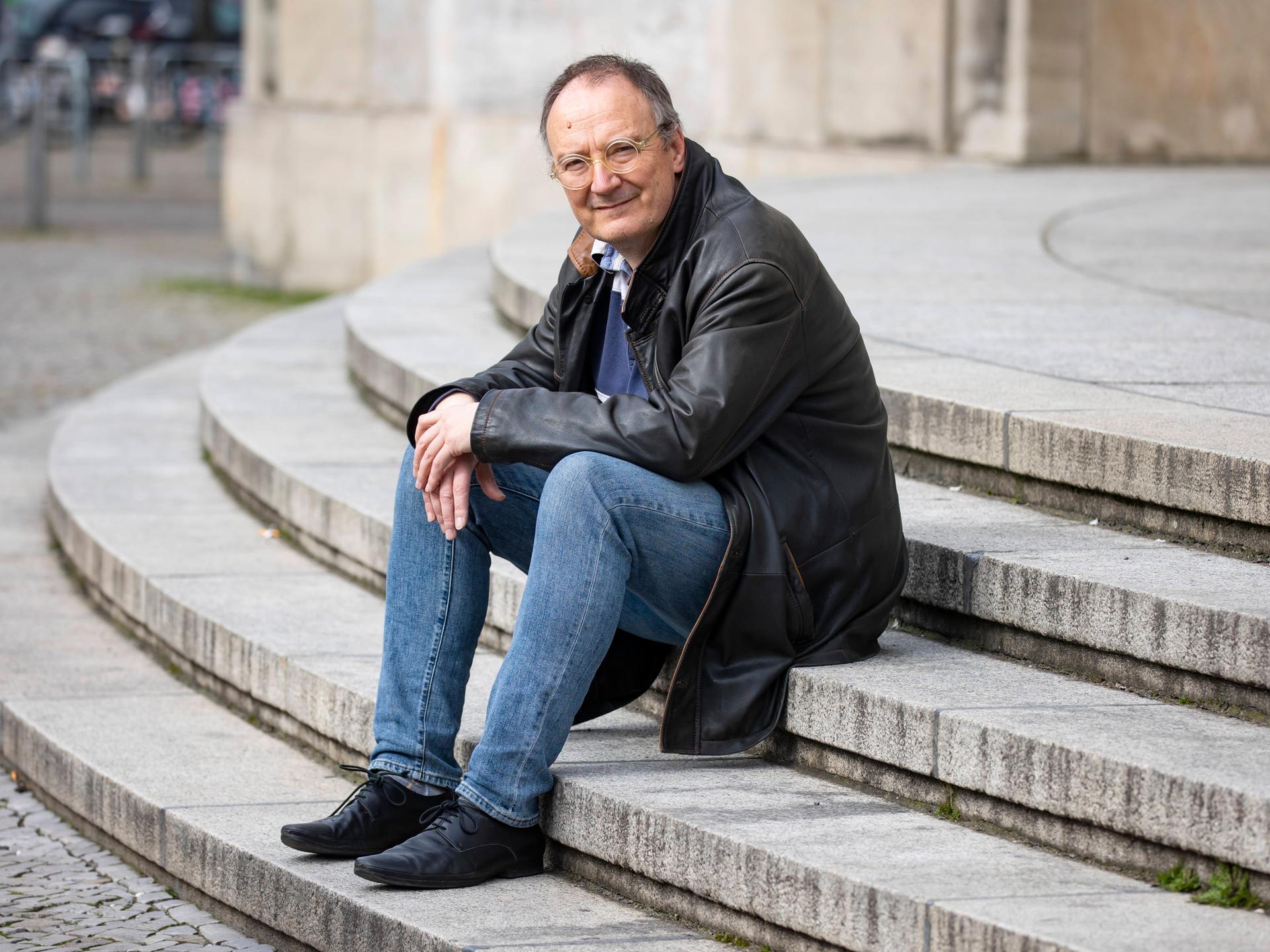 Thomas Brussig sitzt in Lederjacke und Jeans lässig auf den Stufen vor der Berliner Volksbühne und blickt in die Kamera.