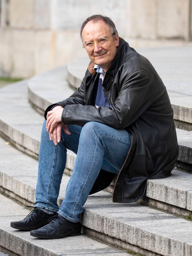 Thomas Brussig sitzt in Lederjacke und Jeans lässig auf den Stufen vor der Berliner Volksbühne und blickt in die Kamera.