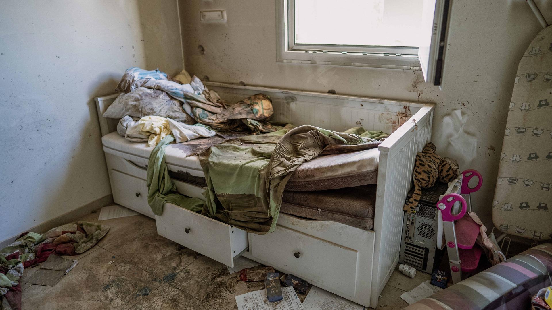 Das Bild zeigt ein zerstörtes Bett in einem Wohnraum. Die Matratze ist herausgerissen und verschmutzt, die Schubladen halb geöffnet. An der Wand sind Einschusslöcher zu sehen.  