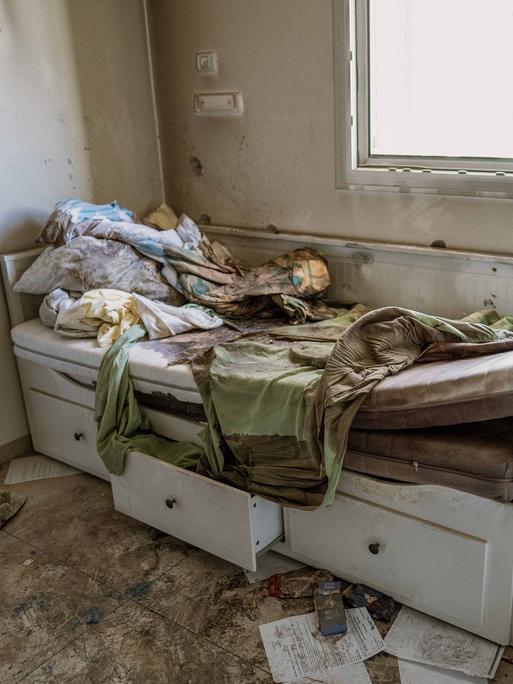 Das Bild zeigt ein zerstörtes Bett in einem Wohnraum. Die Matratze ist herausgerissen und verschmutzt, die Schubladen halb geöffnet. An der Wand sind Einschusslöcher zu sehen.  