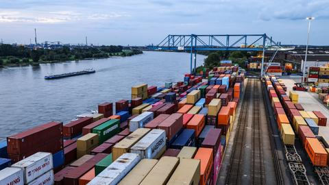 Container stehen im Duisburger Hafen am Kai: Im Containerterminal Logport 2 werden Güter von der Schiene auf Binnenschiffe umgeschlagen. Links im Bild ist ein Fluss zu sehen, auf dem ein Schiff fährt.