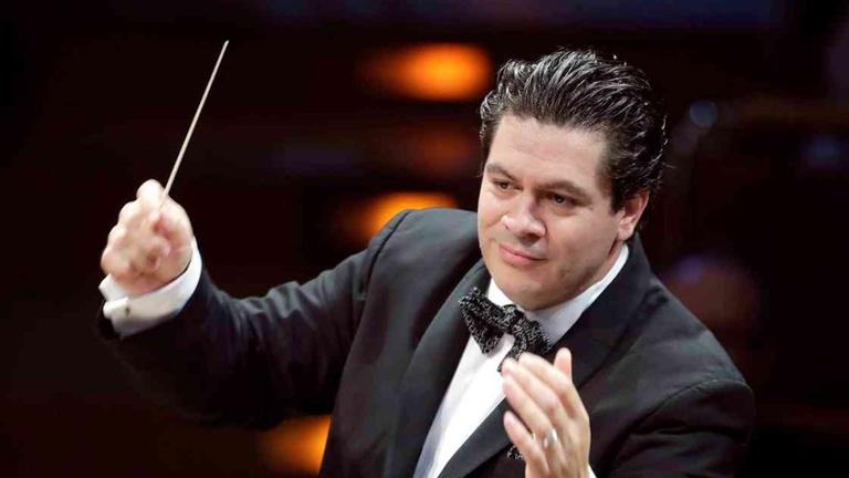 Ein Mann mit schwarzen Haaren und schwarzem Anzug hat die Hände erhoben. In der rechten Hand hält er einen Dirigentenstab.