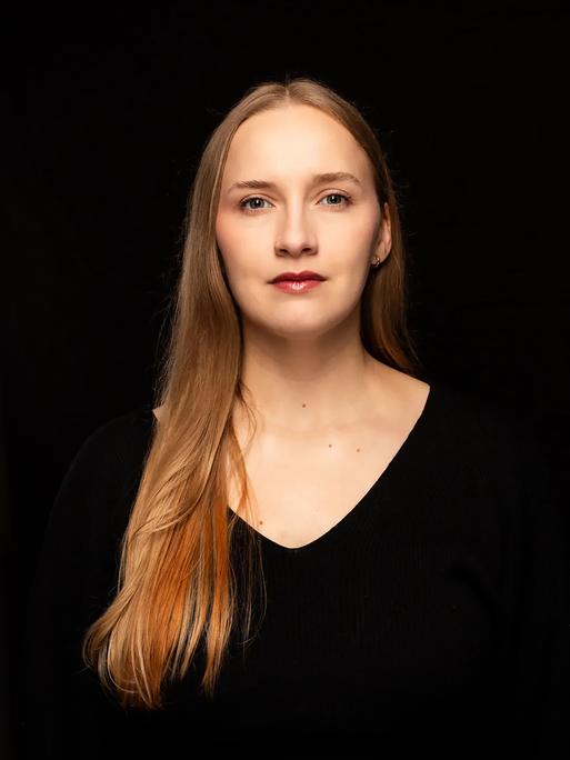 Leonie Plaar trägt ein schwarzes Oberteil und steht vor einem schwarzen Hintergrund. Sie schaut direkt in die Kamera. 