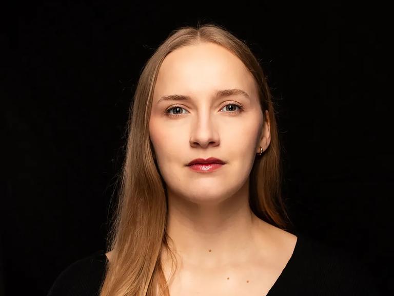 Leonie Plaar trägt ein schwarzes Oberteil und steht vor einem schwarzen Hintergrund. Sie schaut direkt in die Kamera. 