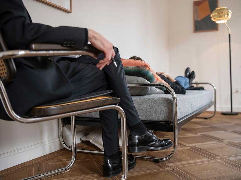 Ein Psychotherapeut, sitzend, therapiert in seiner Praxis eine Klientin, die auf einer Tagesbett liegt.