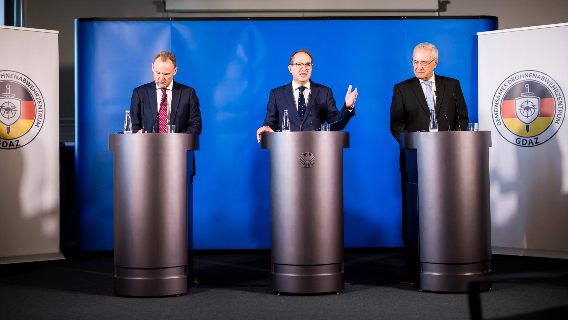 Pressekonferenz zur Eröffnung des Gemeinsamen Drohnenabwehrzentrums. Drei Männer in dunklen Anzügen stehen an Rednerpulten, der Mittlere spricht und gestikuliert.