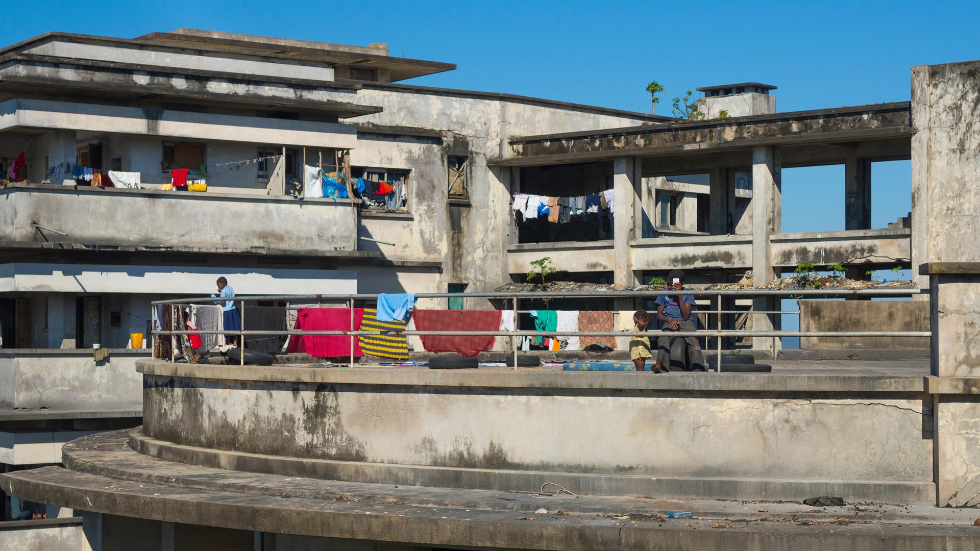 Blick auf die Ruine eines Hotels, auf dessen Balkonen Menschen zu sehen sind und auf dessen Brüstungen bunte Handtücher hängen. Blick auf die Ruine eines Hotels, auf dessen Balkonen Menschen zu sehen sind und auf dessen Brüstungen bunte Handtücher hängen.