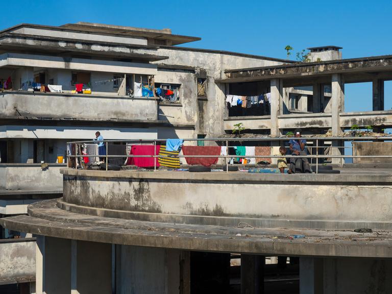 Blick auf die Ruine eines Hotels, auf dessen Balkonen Menschen zu sehen sind und auf dessen Brüstungen bunte Handtücher hängen.