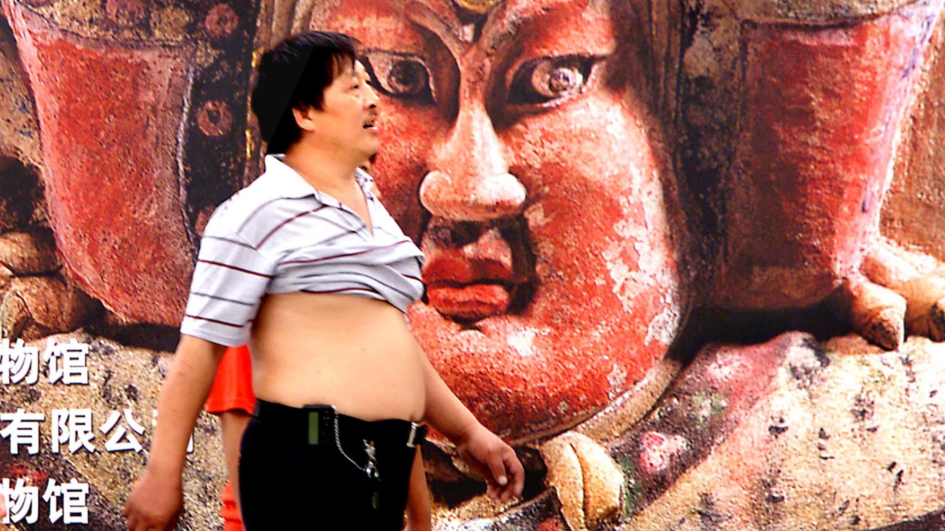 Ein Mann bis unter die Achseln hochgerolltem T-Shirt laüft in Peking an einem Werbeplakat vorbei.