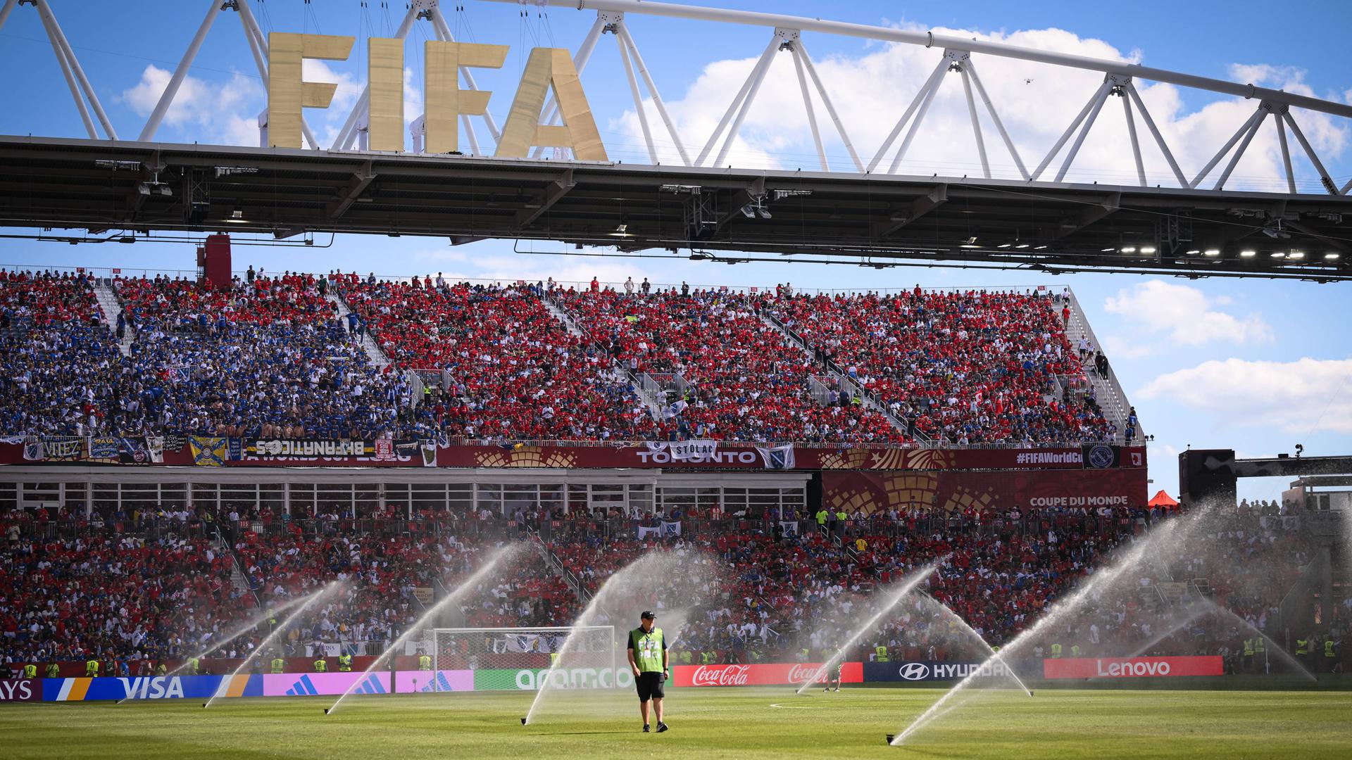 Ein Mensch steht im Fußball-Stadion auf dem Rasen. Der Rasen wird gerade bewässert. Im Hintergrund sind die Ränge mit vielen Fans und ein großes Schild mit der Aufschrift "FIFA". Ein Mensch steht im Fußball-Stadion auf dem Rasen. Der Rasen wird gerade bewässert. Im Hintergrund sind die Ränge mit vielen Fans und ein großes Schild mit der Aufschrift "FIFA".