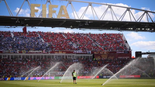 Ein Mensch steht im Fußball-Stadion auf dem Rasen. Der Rasen wird gerade bewässert. Im Hintergrund sind die Ränge mit vielen Fans und ein großes Schild mit der Aufschrift "FIFA".