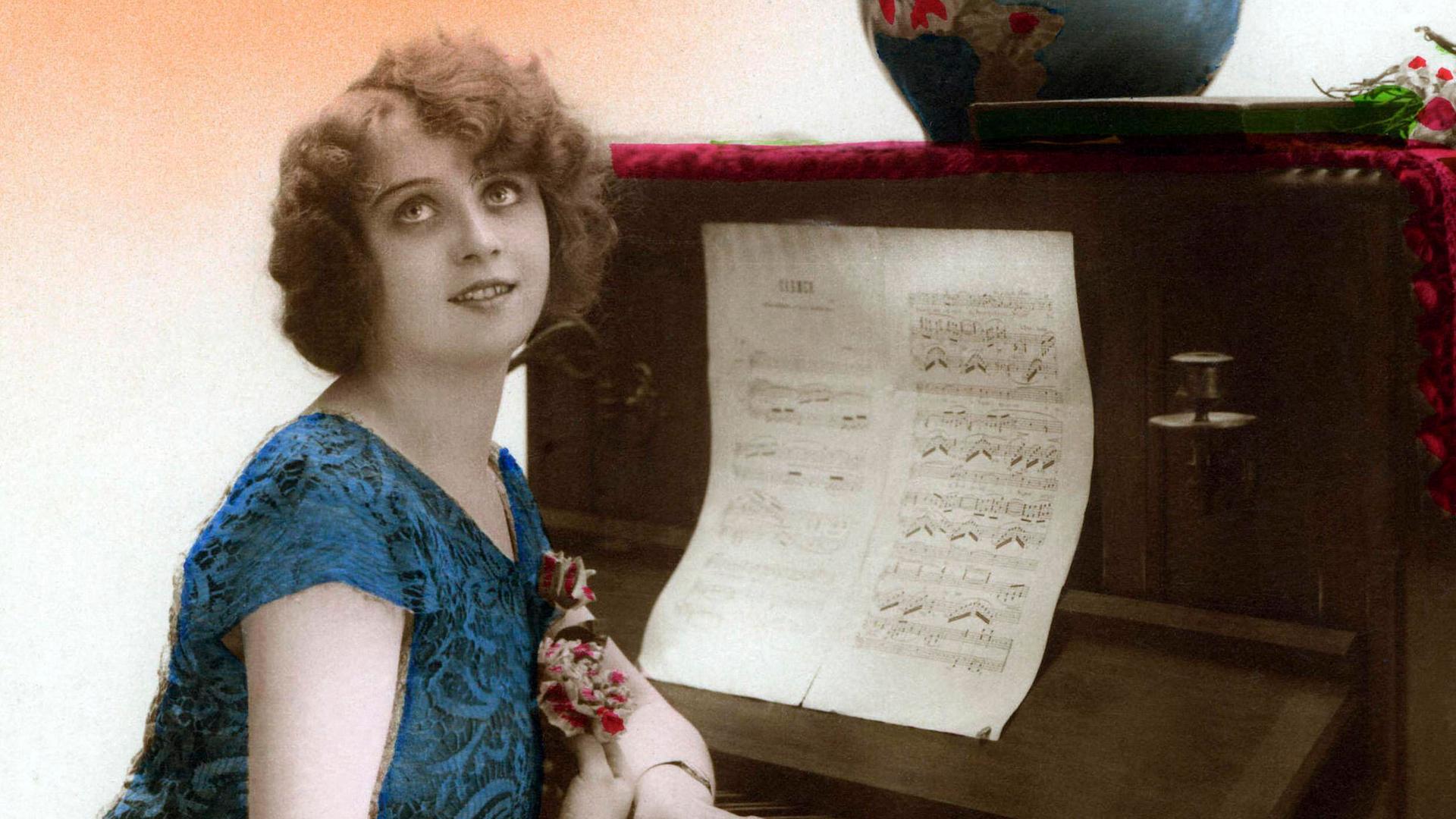 Historische Fotographie um 1905 einer Frau, die am Klavier sitzt und dabei verklärt die Augen nach oben richtet.