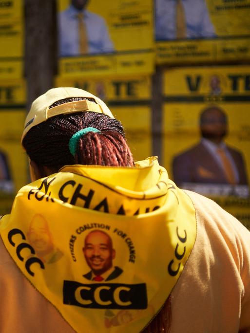 Ein Unterstützer der Oppositionspartei trägt eine gelbe Jacke mit einem aufgedruckten Porträt des Kandidaten. Im Hintergrund sind verschwommen Wahlplakate zu sehen.
