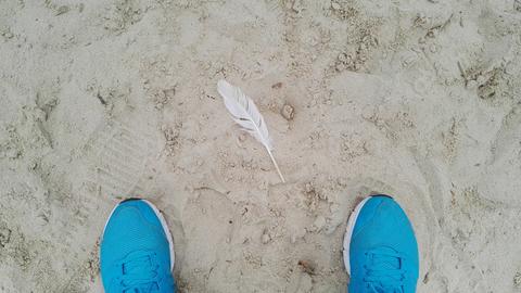 Vogelperspektive auf eine im Sand liegende Feder. Links und rechts im unteren Bildrand die Spitzen blauer Turnschuhe.