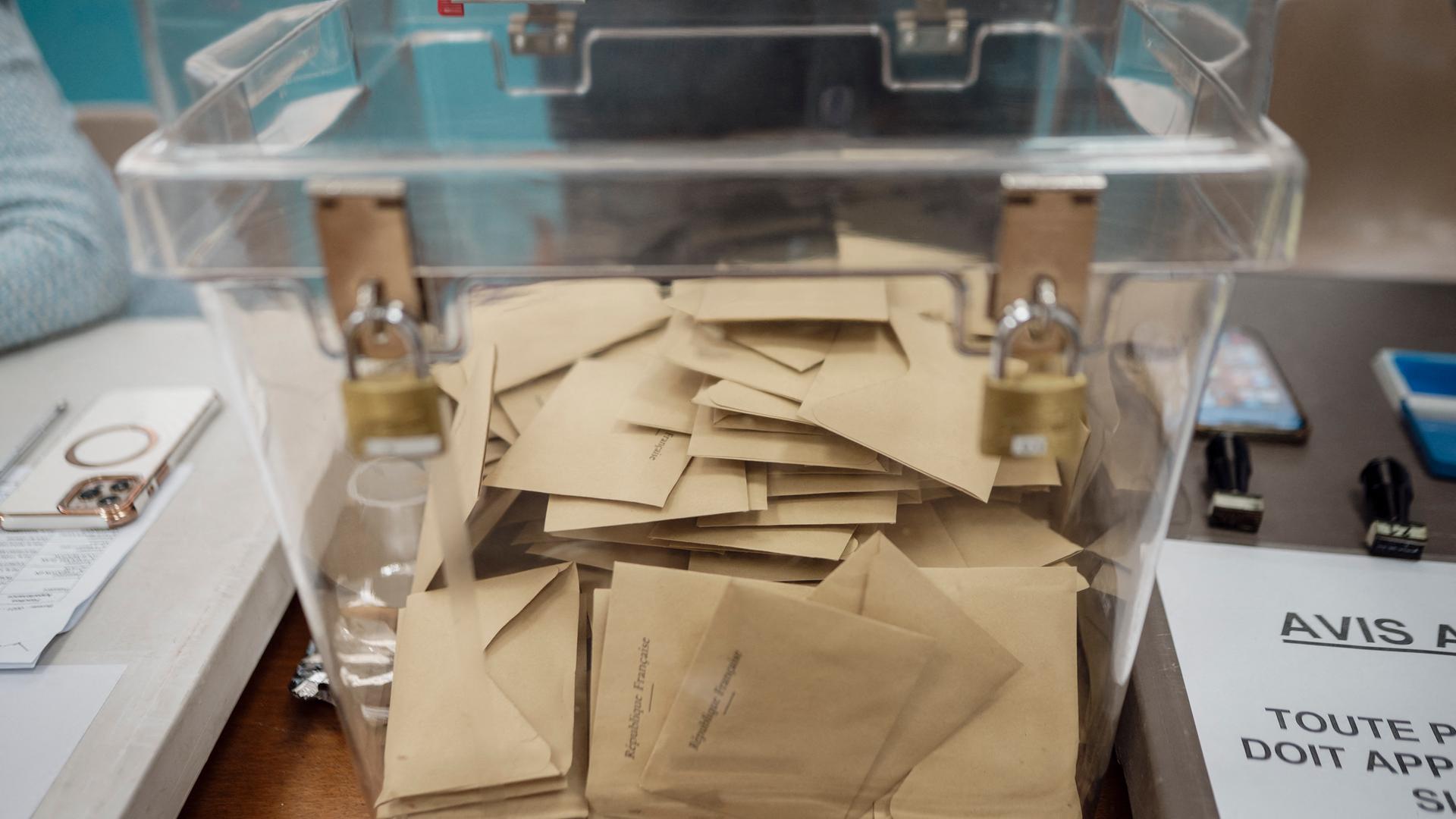 In der auf einem Tisch stehenden durchsichtigen Urne liegen viele braune Umschläge. An einer Seite der Urne befinden sich zwei Schlösser, mit denen sie verschlossen werden.