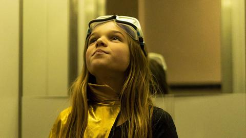 Ein Mädchen im Superheldinnenkostüm steht in einem Aufzug und blickt nach oben.