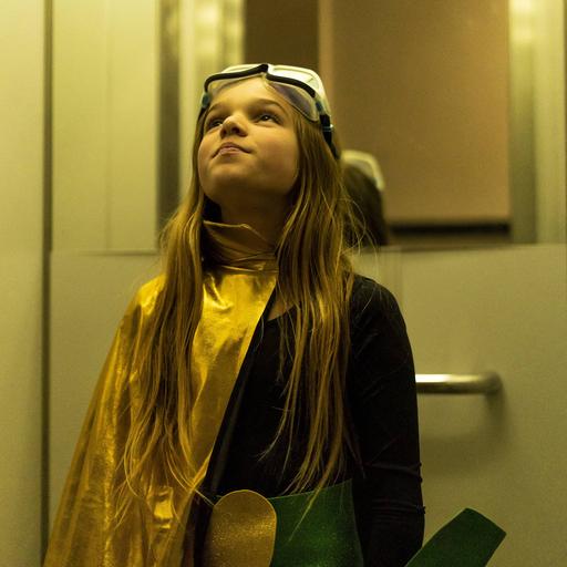 Ein Mädchen im Superheldinnenkostüm steht in einem Aufzug und blickt nach oben.