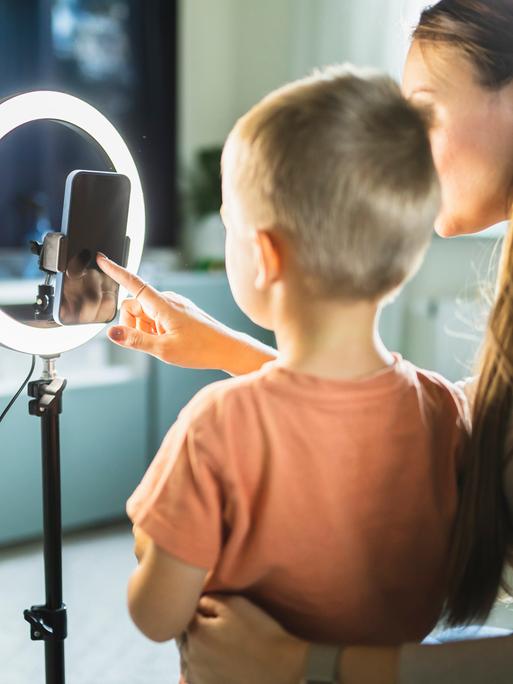Eine Mutter und ein Kind sitzen mit einem Smartphone vor einem Ringlicht und sprechen in die Kamera des Telefons. (Symbolbild)