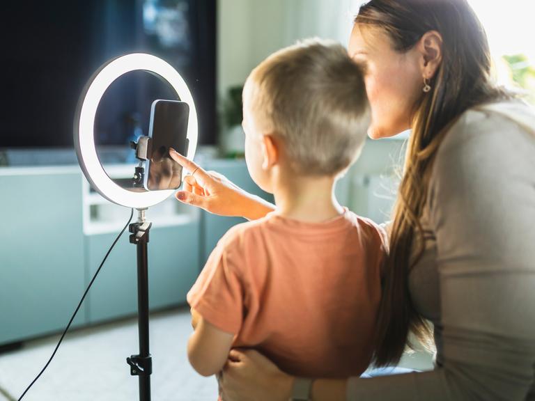 Eine Mutter und ein Kind sitzen mit einem Smartphone vor einem Ringlicht und sprechen in die Kamera des Telefons. (Symbolbild)