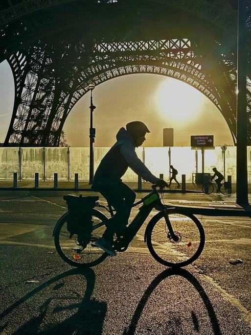 Ein Radfahrer mit Helm fährt nachts im Gegenlicht vor der Stahlkonstruktion des Eiffelturms vorbei. 