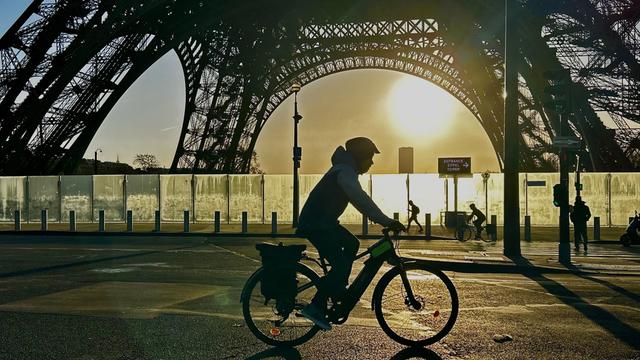 Ein Radfahrer mit Helm fährt nachts im Gegenlicht vor der Stahlkonstruktion des Eiffelturms vorbei. 