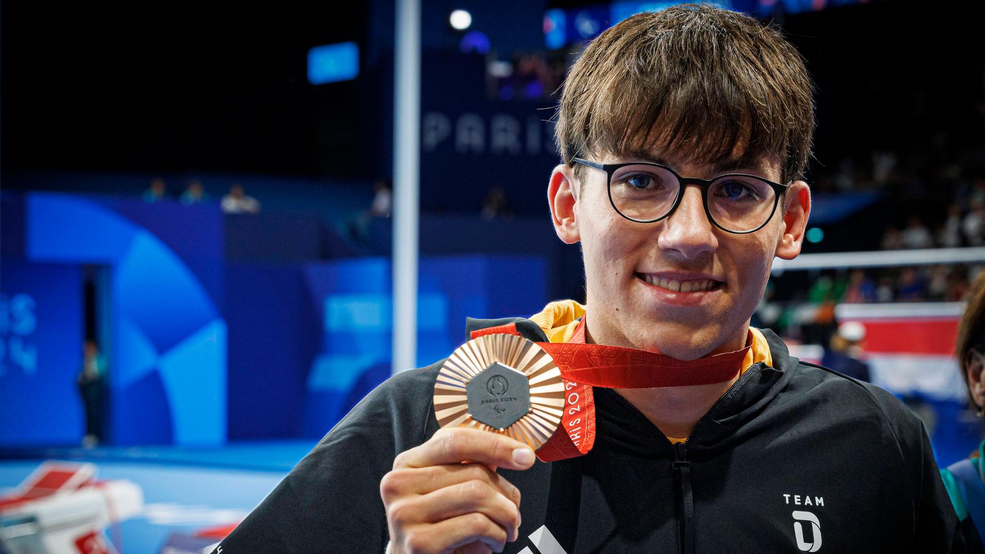 Maurice Wetekam zeigt seine Bronzemedaille und freut sich. Er steht in der Schwimm-Arena in Paris und trägt einen schwarzen Kapuzenpullover.