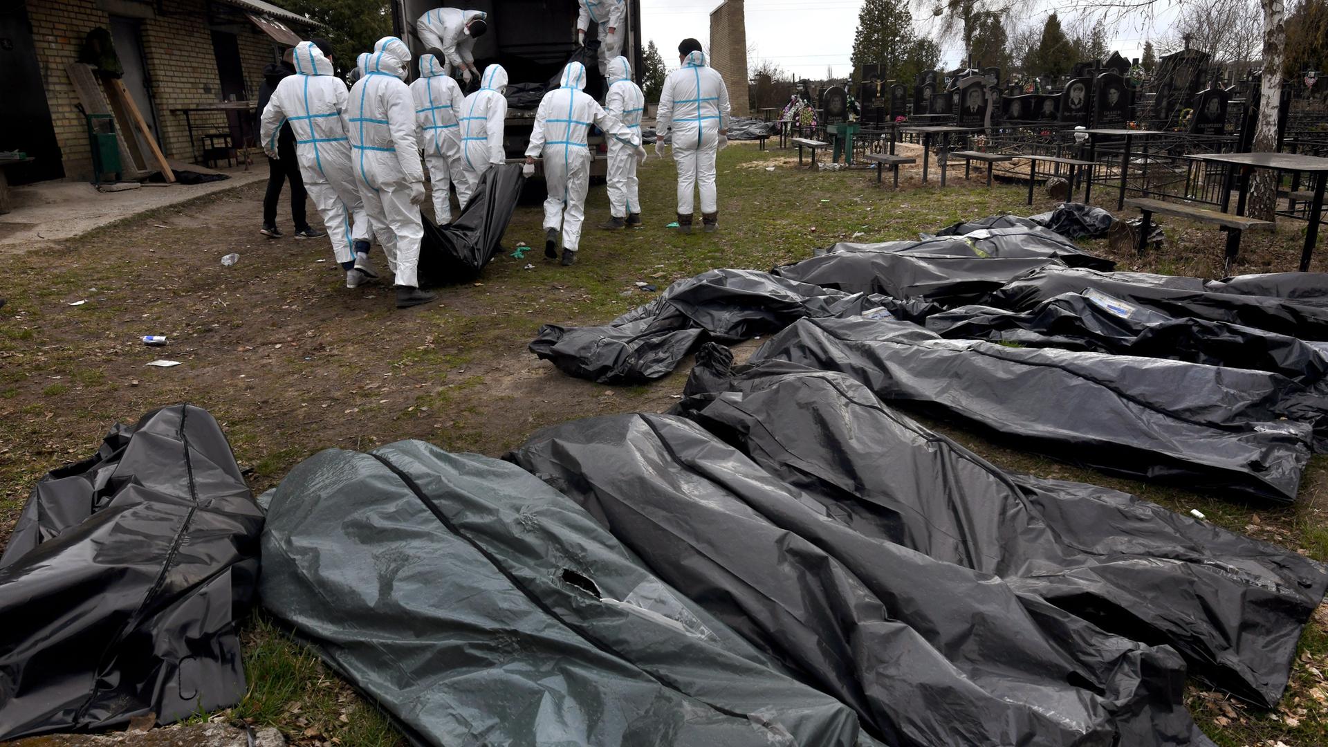 Mehrere Leichensäcke aus Plastik liegen in einer Reihe, im Hintergrund tragen mehrere Personen in weißen Schutzanzügen die Leichensäcke zu einem Lastwagen. 