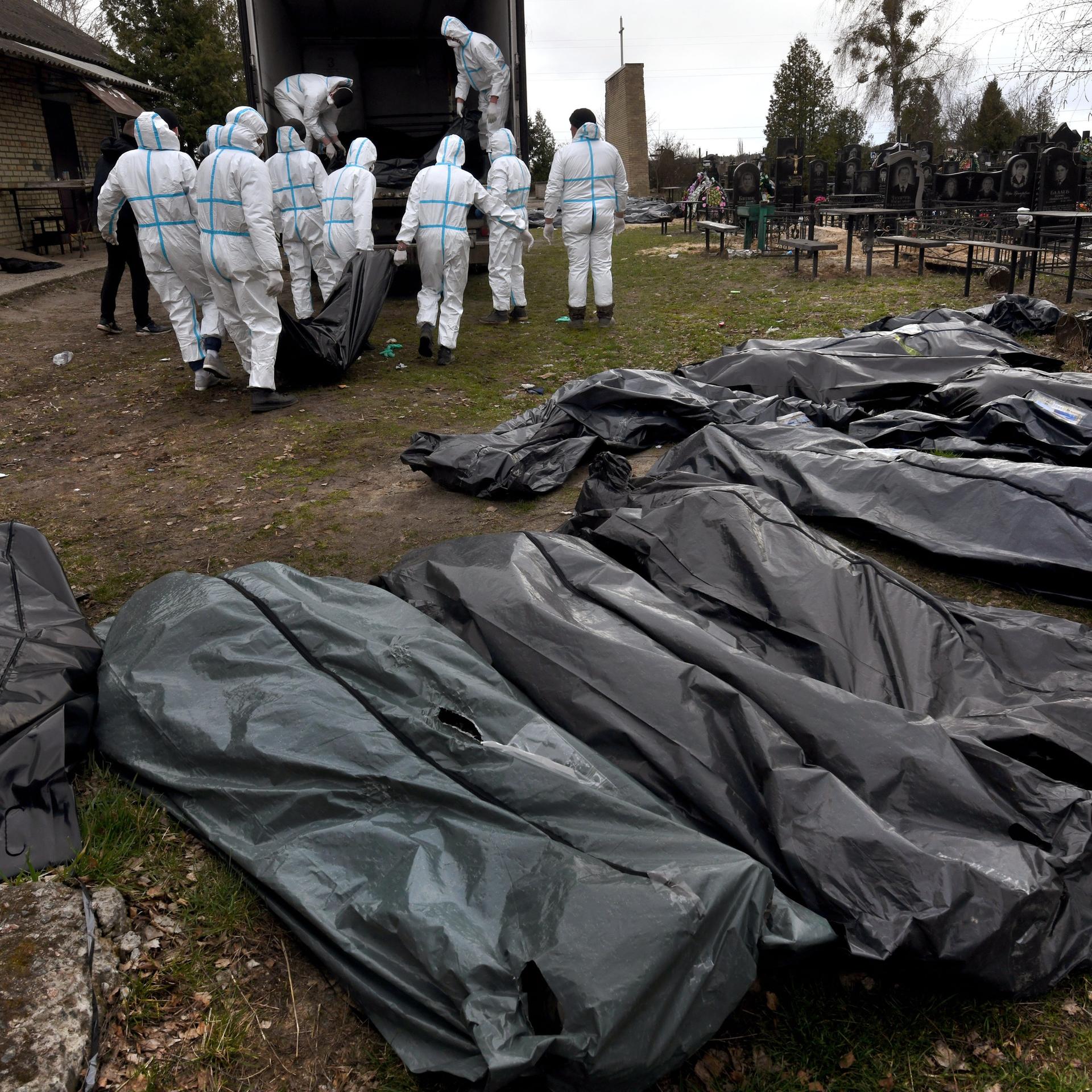 Mehrere Leichensäche aus Plastik liegen in einer Reihe, im Hintergrund tragen mehrere Personen in weißen Schutzanzügen die Leichensäcke zu einem Lastwagen. 