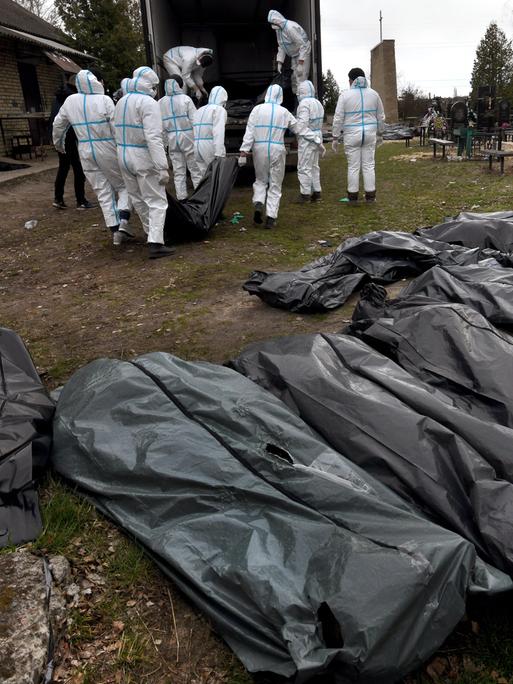 Mehrere Leichensäcke aus Plastik liegen in einer Reihe, im Hintergrund tragen mehrere Personen in weißen Schutzanzügen die Leichensäcke zu einem Lastwagen. 