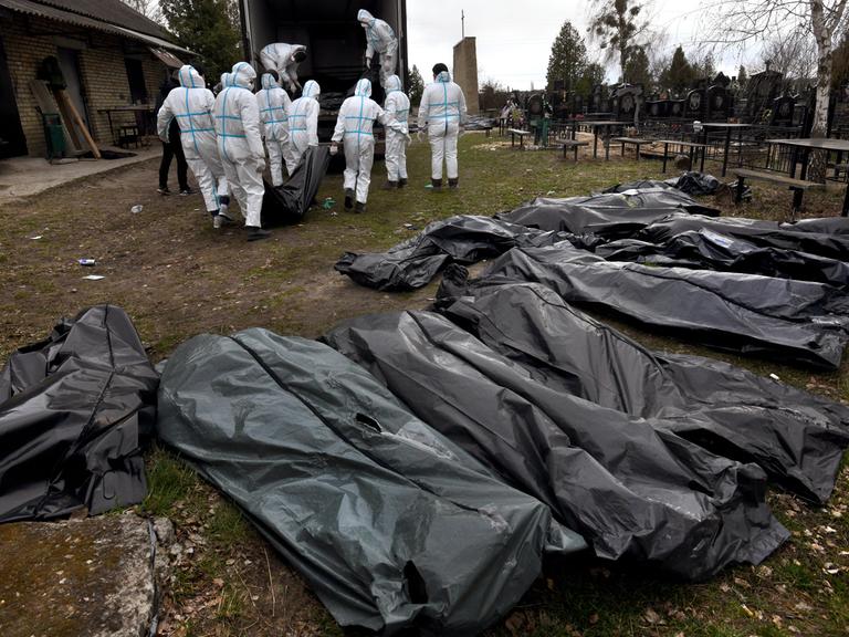 Mehrere Leichensäcke aus Plastik liegen in einer Reihe, im Hintergrund tragen mehrere Personen in weißen Schutzanzügen die Leichensäcke zu einem Lastwagen. 