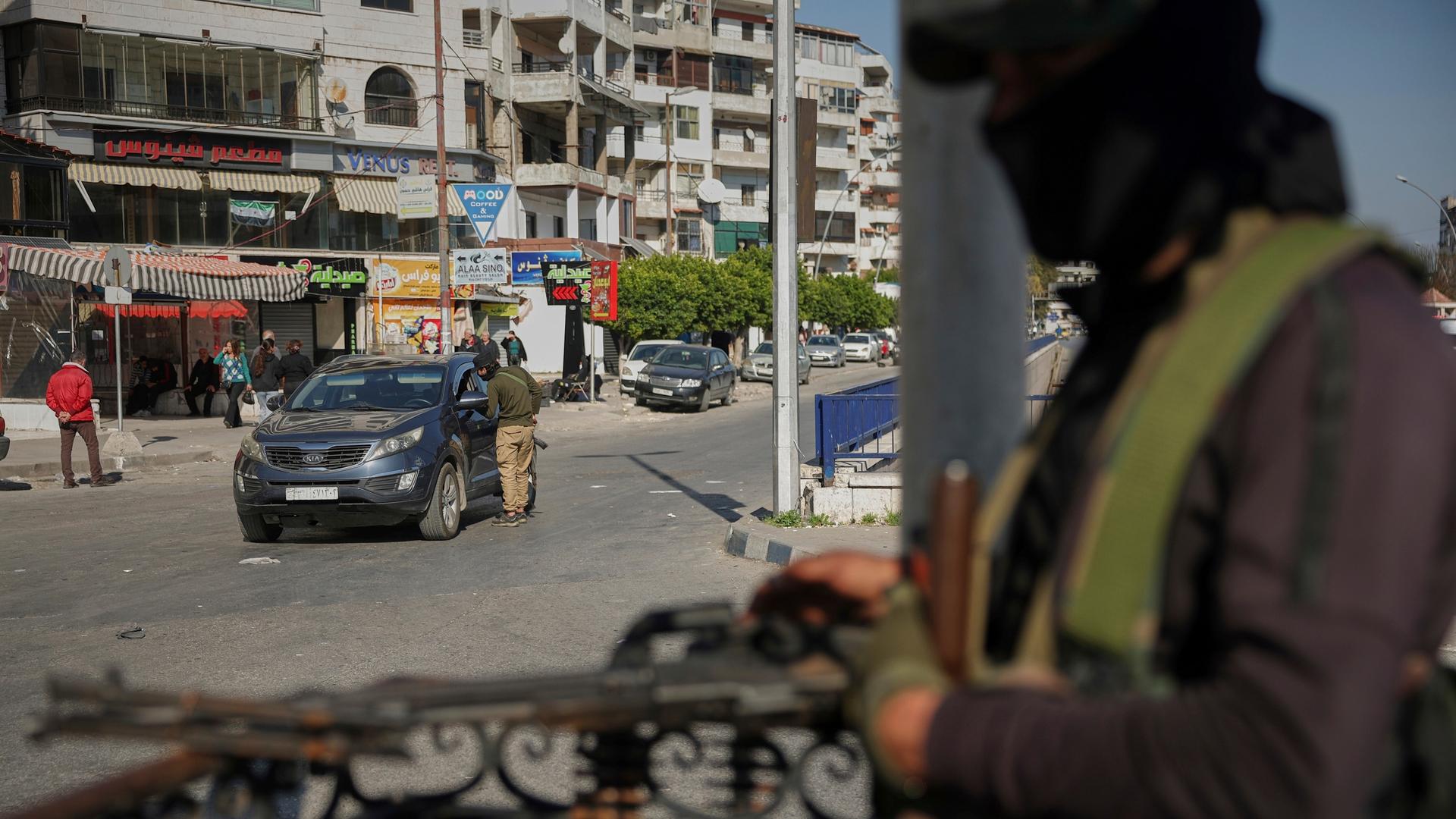 Im Vordergrund steht ein bewaffneter Mann mit Sturmhaube, im Hintergrund ist zu sehen, wie ein Sicherheitsmann einen Autofahrer kontrolliert. Im Vordergrund steht ein bewaffneter Mann mit Sturmhaube, im Hintergrund ist zu sehen, wie ein Sicherheitsmann einen Autofahrer kontrolliert.